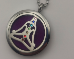 Colgante aromático de acero inoxidable con un diseño de una mujer en pose de yoga, incrustado con piedras de colores alineadas con los siete chakras sobre un fondo morado, ideal para usar con aceites esenciales y promover el equilibrio espiritual.