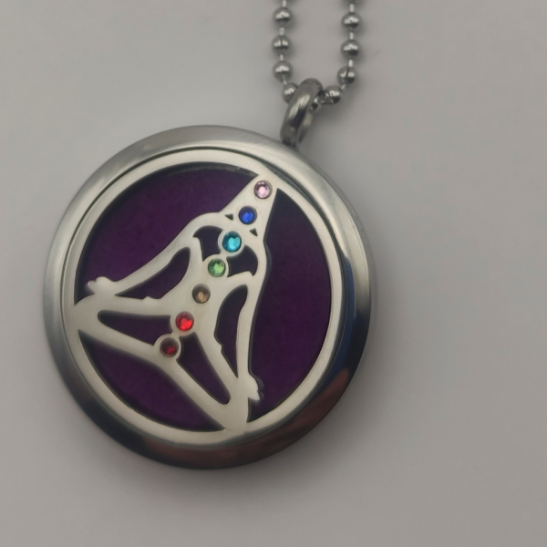 Colgante aromático de acero inoxidable con un diseño de una mujer en pose de yoga, incrustado con piedras de colores alineadas con los siete chakras sobre un fondo morado, ideal para usar con aceites esenciales y promover el equilibrio espiritual.
