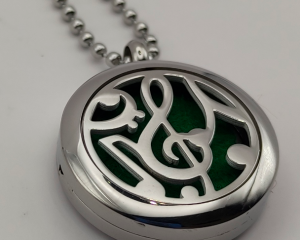 Colgante aromático de acero inoxidable con un diseño de clave de sol y notas musicales en la tapa sobre un fondo verde, ideal para usar con aceites esenciales y combinar estilo musical con bienestar.