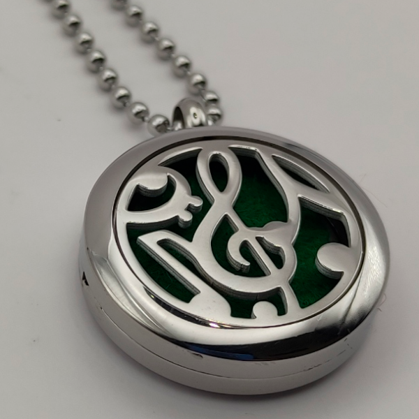 Colgante aromático de acero inoxidable con un diseño de clave de sol y notas musicales en la tapa sobre un fondo verde, ideal para usar con aceites esenciales y combinar estilo musical con bienestar.